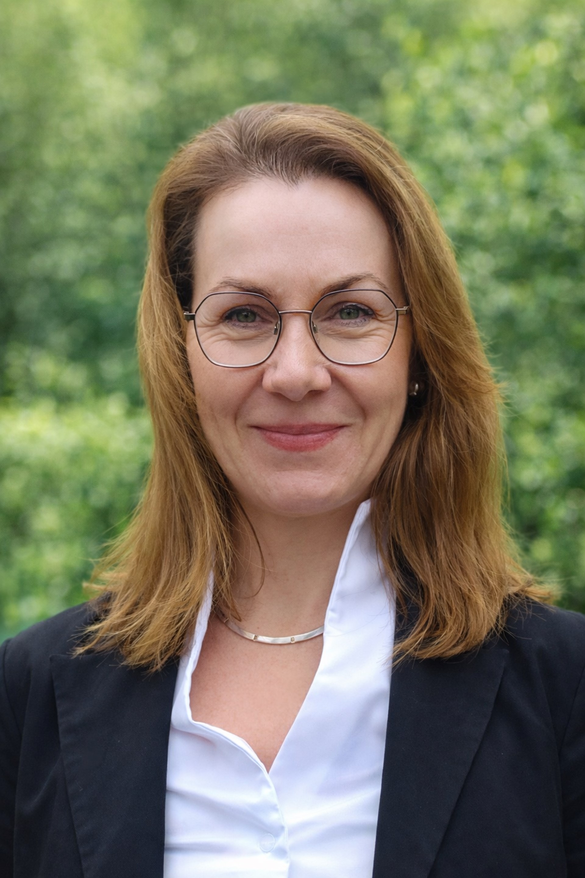 Porträt einer lächelnden Frau mit schulterlangen hellbraunen Haaren und runder Brille. Sie trägt eine weiße Bluse, ein dunkles Sakko und eine schlichte Halskette. Der Hintergrund ist unscharf und zeigt grünes Laub, wodurch die Person klar im Vordergrund steht.